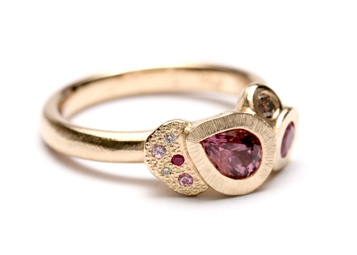 A gold ring featuring a teardrop-shaped pink gemstone centerpiece, surrounded by small pink and clear stones on a textured gold setting, with a polished gold band, shown on a white background.